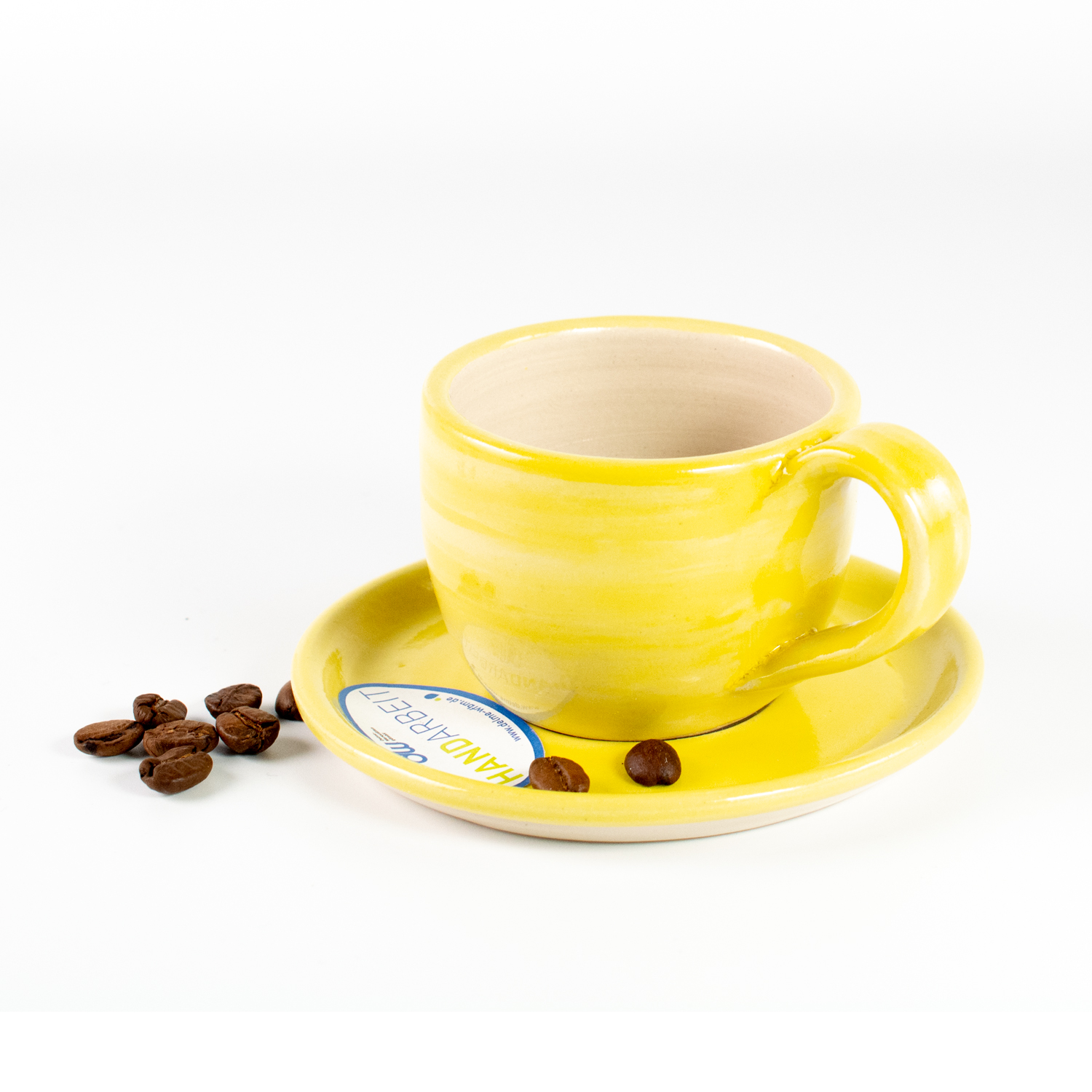 Eine gelbe leicht durchscheinend glasierte Cappuccino Tasse auf einer Untertasse, mit gleichfarbigem Rand. Das innere der Tasse und Untertasse ist in ein naturbelassenes helles Beige. Daneben liegen ein paar Kaffeebohnen.