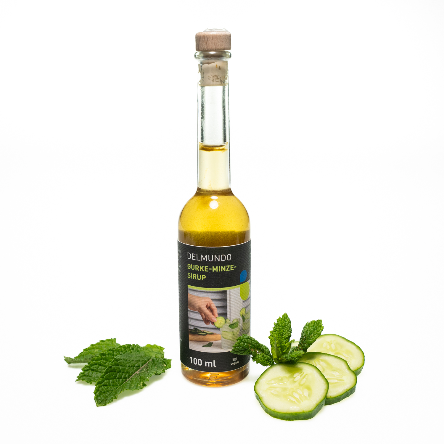Eine klare Glasflasche mit Bügel-Verschluss gefüllt mit erfrischend grün-goldenem Gurke-Minz-Sirup. Daneben liegen Gurkenscheiben und Minzblätter.
